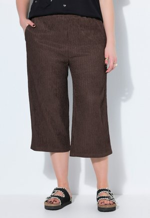 Dolna część ciała osoby noszącej brązowe, szerokie, skrócone spodnie i czarne sandały z białą, falistą lamówką, stojącej na tle białej ściany.