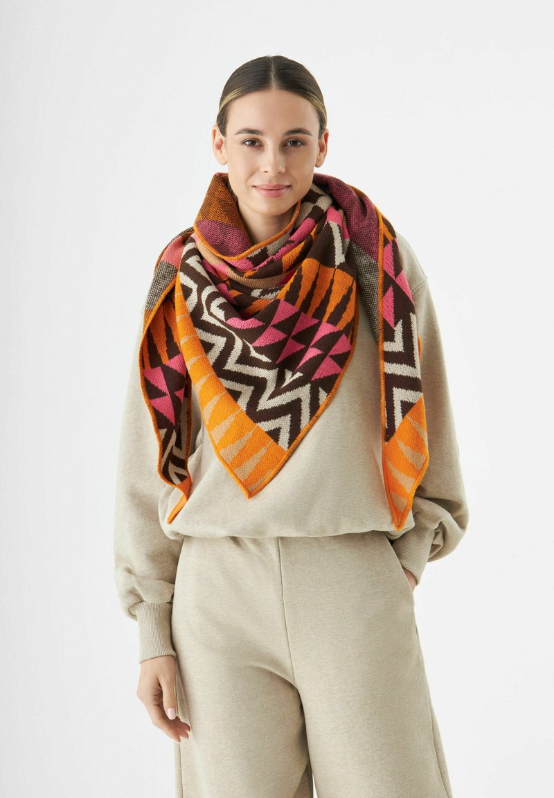 Jong vrouw in een beige sweatshirt en broek met een grote, kleurrijke sjaal met geometrisch patroon in oranje, roze, wit en bruintinten.