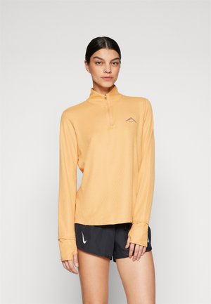 Haut à manches longues avec col zip en orange clair, fabriqué en tissu texturé ; présente un petit logo sur la poitrine et des trous pour les pouces au niveau des poignets.