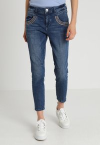 Kvinna i blå denimjeans med dekorativ sömnad på framfickorna och vita snörskor, som står mot en enfärgad bakgrund.