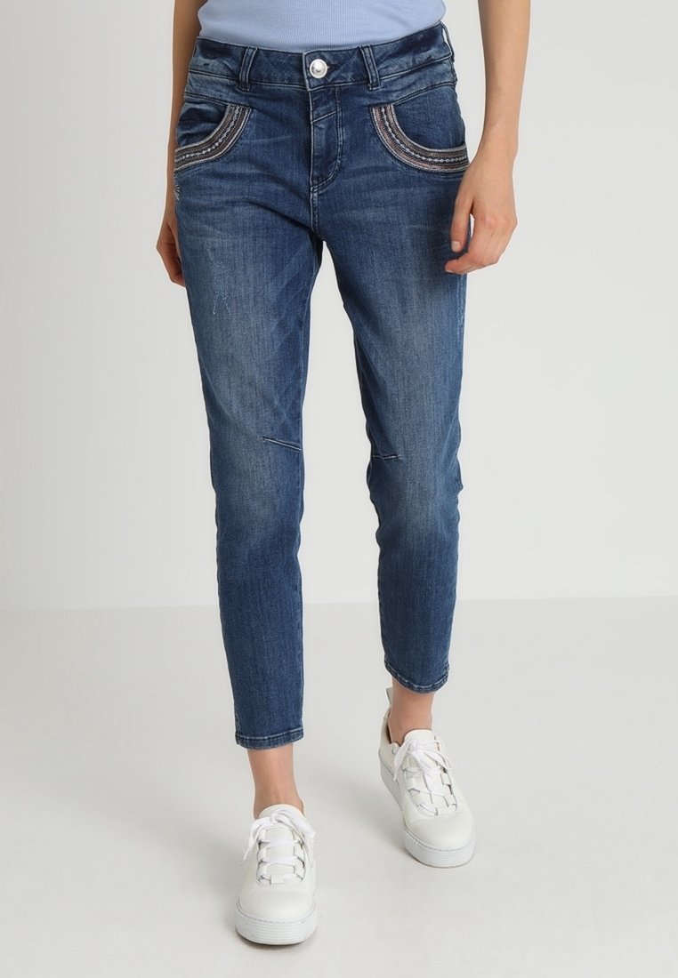 Kvinna i blå denimjeans med dekorativ sömnad på framfickorna och vita snörskor, som står mot en enfärgad bakgrund.