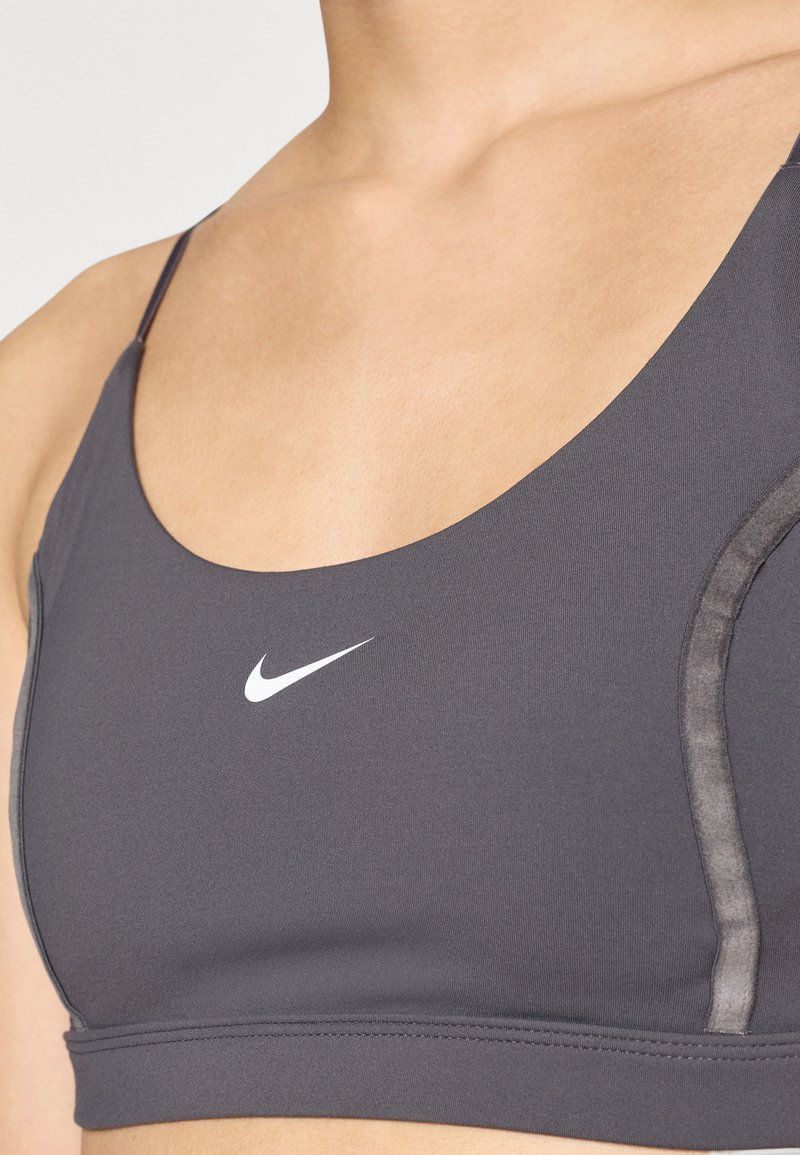 Grijze sportbeha met dunne straps, voorzien van een wit Nike-logo aan de linkerkant. Glad materiaal met subtiele ontwerplijnen langs de zijkanten.