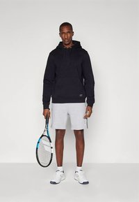 Μαύρο φούτερ με κουκούλα και μπροστινή τσέπη, συνδυασμένο με γκρι σορτς. Κρατάει ρακέτα τένις και φοράει λευκά και μπλε αθλητικά παπούτσια.