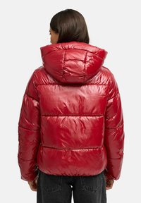 Chaqueta acolchada roja con acabado brillante, capucha y secciones acolchadas horizontales. Cuenta con puños elásticos y un diseño corto.
