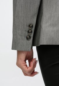 Manche de blazer gris avec trois boutons ronds foncés légèrement brillants, fabriqué à partir d'un tissu texturé, avec une finition nette.