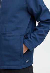 Veste soft-shell bleu marine avec fermeture éclair, poches latérales et finition texturée. Présente un petit logo cousu sur le haut du bras.
