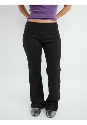 Osoba ubrana w czarne rozszerzane spodnie, fioletowy krótki top i srebrne baletki, stojąca z rękami za plecami na jednolitym tle.