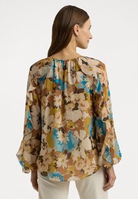 Blomsterbluse i brun, gul og blå, med volanger på ermene og en rund halsutskjæring. Laget av lett, teksturert stoff.