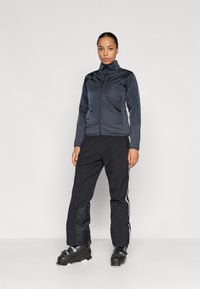Veste zippée gris foncé avec un col haut, associée à un pantalon noir avec des bandes blanches sur les côtés. Portée avec des bottes de ski noires.