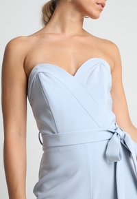 Femme portant une robe sans bretelles de couleur bleu clair avec un décolleté en cœur et une ceinture nouée, se tenant devant un fond uni.
