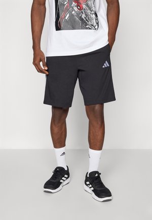 Mężczyzna ubrany w czarne szorty Adidas, białe skarpetki z logo, czarne buty sportowe Adidas i biały t-shirt z grafiką Spider-Mana.