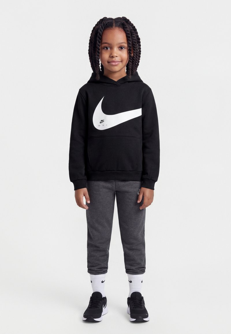 Παιδί με πλεγμένα μαλλιά στέκεται φορώντας μαύρο φούτερ Nike με κουκούλα, σκούρες γκρι φόρμες, λευκές κάλτσες Nike και μαύρα αθλητικά παπούτσια Nike σε λευκό φόντο.