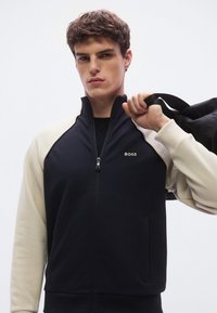 Mustan ja kermanvärisen vetoketjullisen takin korkea kaulus, jossa BOSS-logo. Sileä kangas, moderni leikkaus ja näkyvät sivutaskut.