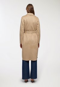 Cappotto trench in suede beige con vita regolabile, maniche lunghe e vestibilità comoda, abbinato a jeans in denim blu a gamba larga e tacchi.