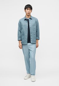 Ανοιχτό μπλε μπουφάν με τέσσερις μπροστινές τσέπες, φορεμένο πάνω από ένα ναυτικό μπλουζάκι. Ανοιχτό μπλε παντελόνι με λεπτές ρίγες και λευκά αθλητικά παπούτσια ολοκληρώνουν την εμφάνιση.