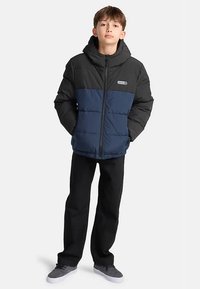 Doudoune avec une section supérieure noire et une section inférieure bleu marine. Dotée d'une capuche, d'une fermeture éclair avant et d'un patch logo. Portée avec un pantalon foncé.