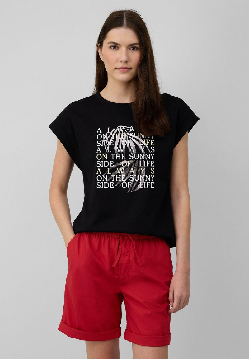 Zwarte katoenen t-shirt met korte mouwen, voorzien van witte tekst en een grafisch ontwerp. Combinatied met rode, opgerolde zomer shorts.