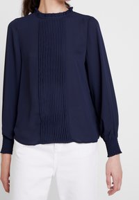 Blouse marine à manches longues, col à volants et plis verticaux sur le devant. Fabriquée en tissu lisse avec des poignets froncés.