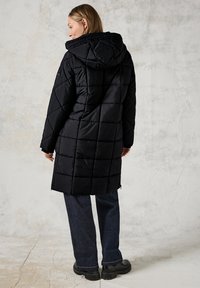 Svart vadderad långjacka med huva, har en quiltad design, texturerat tyg och en avslappnad passform. Kombinerad med mörka denimbyxor och stövlar.
