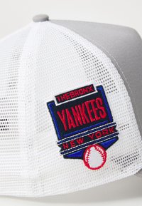Grå cap med hvit netting bak. Har et fargerikt brodert Yankees-logo, med teksten "THE BRONX" og "NEW YORK", og et baseballgrafikk.