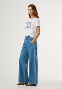 T-shirt bianca con la scritta "PEPE JEANS" stampata in lettere colorate, abbinata a jeans blu a gamba larga. Il modello indossa scarpe beige a punta.