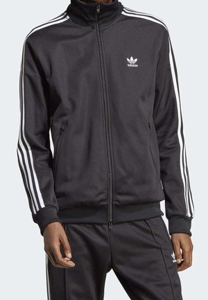 Man som bär en svart Adidas-träningsoverall med vita ränder på ärmarna och byxorna, stående med avslappnade händer vid sidorna.