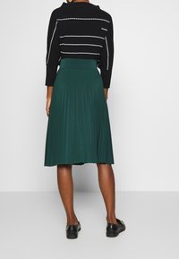 Grüner plissierter Rock mit einem taillierten Bund, kombiniert mit einem schwarzen gestreiften Pullover. Schwarze flache Schuhe vervollständigen das Outfit.