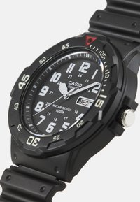 Montre quartz Casio avec bracelet en plastique noir, cadran rond noir, marqueurs horaires blancs, affichage de la date et un accent rouge à 12 heures. Résistante à l'eau jusqu'à 100 mètres.