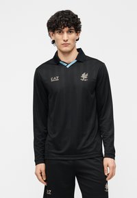 Camiseta de fútbol negra de manga larga con un cuello azul claro, que presenta un logo dorado en el pecho y tela texturizada. Combinada con pantalones negros.