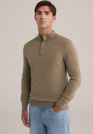 Pullover marrón de manga larga, hecho de tejido suave, con un cuello alto y una tapeta con botones. Lleva puesto sobre unos jeans de color azul claro.