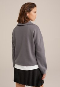 Grå sweatshirt med krave, blød tekstur, afslappet pasform og ballonærmer, sat sammen med en plisseret sort nederdel over en hvid skjorte.