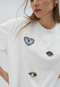 Camiseta blanca de algodón con múltiples adornos en forma de ojo, adornada con cuentas de colores y strass en varias formas y tamaños.
