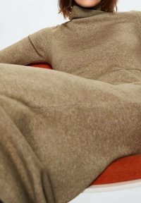 Suéter de cuello alto largo y ajustado en suave tela de punto marrón. Presenta un cuello alto y una textura suave, usado mientras está sentado. El color de fondo es claro.