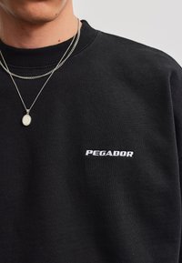 Persona que lleva una sudadera negra con el texto "PEGADOR" en el pecho y dos collares de cadena plateados, uno con un colgante redondo.