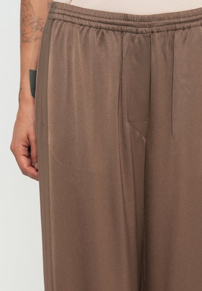 Close-up of brown elastic waist pants worn by a person with tattooed arm resting beside the leg against a plain background.