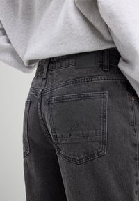 Musta farkkushortsit korkealla vyötäröllä, joissa on takataskut ja teksturoitu pinta. Mukana on brändilogo vyötärönauhassa.