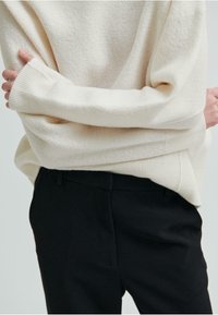 Maglione color crema con un tessuto morbido e texturizzato e polsini a coste, abbinato a pantaloni neri su misura, che mostrano un fit rilassato.