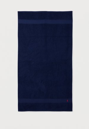 Toalla rectangular azul marino oscuro con dos bandas horizontales cerca de los bordes y un pequeño logo rojo bordado en la esquina inferior derecha.