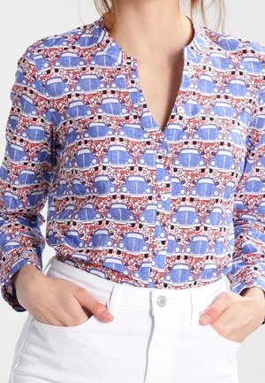 Lange mouwen shirt met een print van een blauwe vintage auto en witte sterren, ingestopt in een wit broek met hoge taille, handen in de zakken.