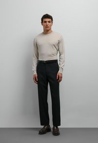 Chemise beige à manches longues, pantalons foncés, ceinture marron et mocassins. Coupe ajustée, texture lisse, design minimaliste sur un fond gris clair.