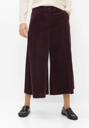 Vrouw draagt een wijd-uitlopende, cropped donkerbruine corduroy broek, een crème trui en zwarte leren loafers, staand tegen een witte achtergrond.