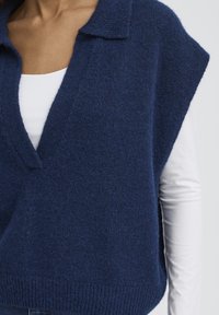 Veston en tricot bleu marine avec col en V, col côtelé et larges emmanchures, superposé sur un haut blanc à manches longues. Tissu à finition texturée.