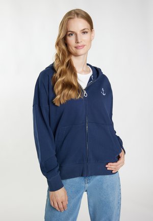 Woman with long wavy blonde hair wearing a navy blue zip-up hoodie with anchor logo and light blue jeans, standing against a plain background.