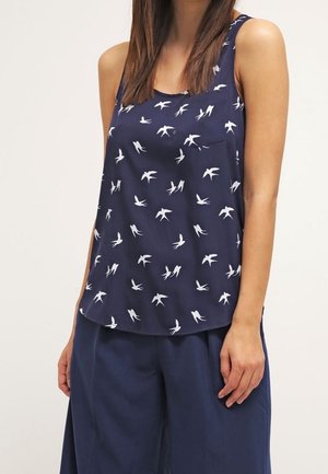 Femme portant un haut sans manches bleu marine avec des motifs d'oiseaux blancs et un pantalon large bleu marine sur un fond clair uni.
