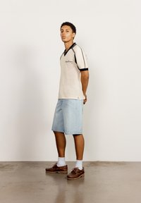Ung kvinne står sidelengs mot en hvit vegg iført en beige strikket kortermet skjorte, lyse denimshorts, hvite sokker og brune sko.