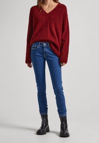 Suéter rojo, de gran tamaño, tejido a mano con escote en V; combinado con jeans oscuros de corte ajustado y botas negras de tacón grueso.