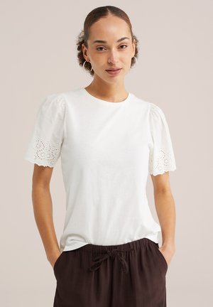 Donna con capelli ricci tirati indietro che indossa orecchini a cerchio, una camicetta bianca a maniche corte con dettagli a traforo e pantaloni scuri con coulisse.