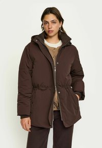 Bruine pufferjack met een capuchon, een trekkoord in de taille, een rits aan de voorkant en drukknopen, gedragen over een beige overhemd met knopen. Gladde textuur.