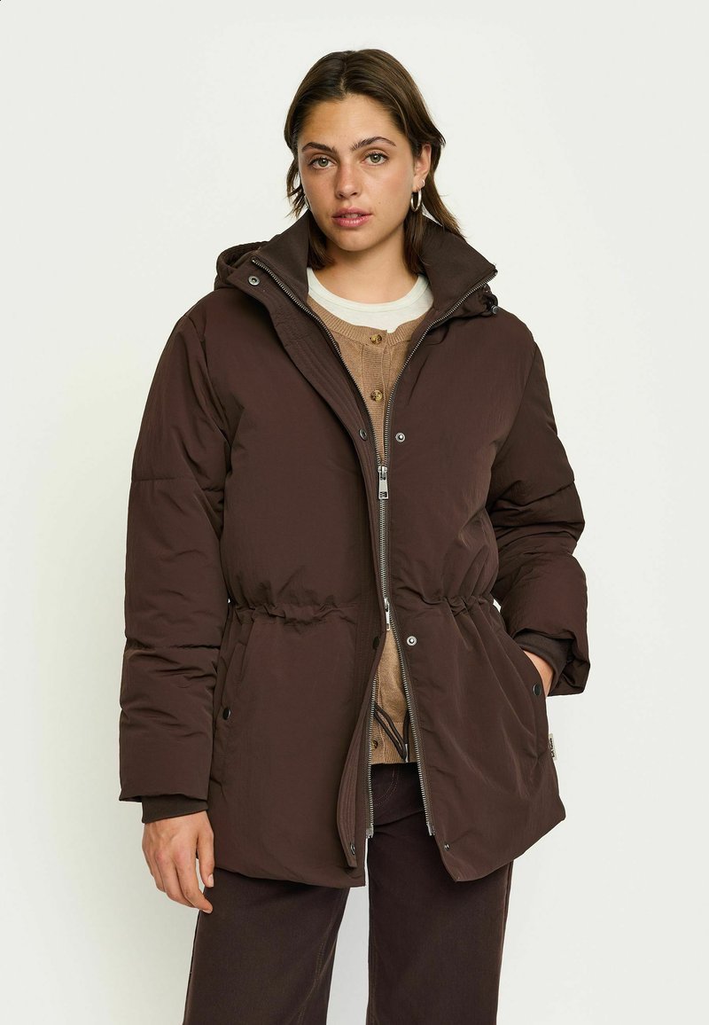 Bruine pufferjack met een capuchon, een trekkoord in de taille, een rits aan de voorkant en drukknopen, gedragen over een beige overhemd met knopen. Gladde textuur.