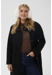 Femme aux longs cheveux blonds portant des cache-oreilles blancs pelucheux, un manteau noir texturé, un pull marron et un jean bleu foncé, souriant avec une main dans la poche.